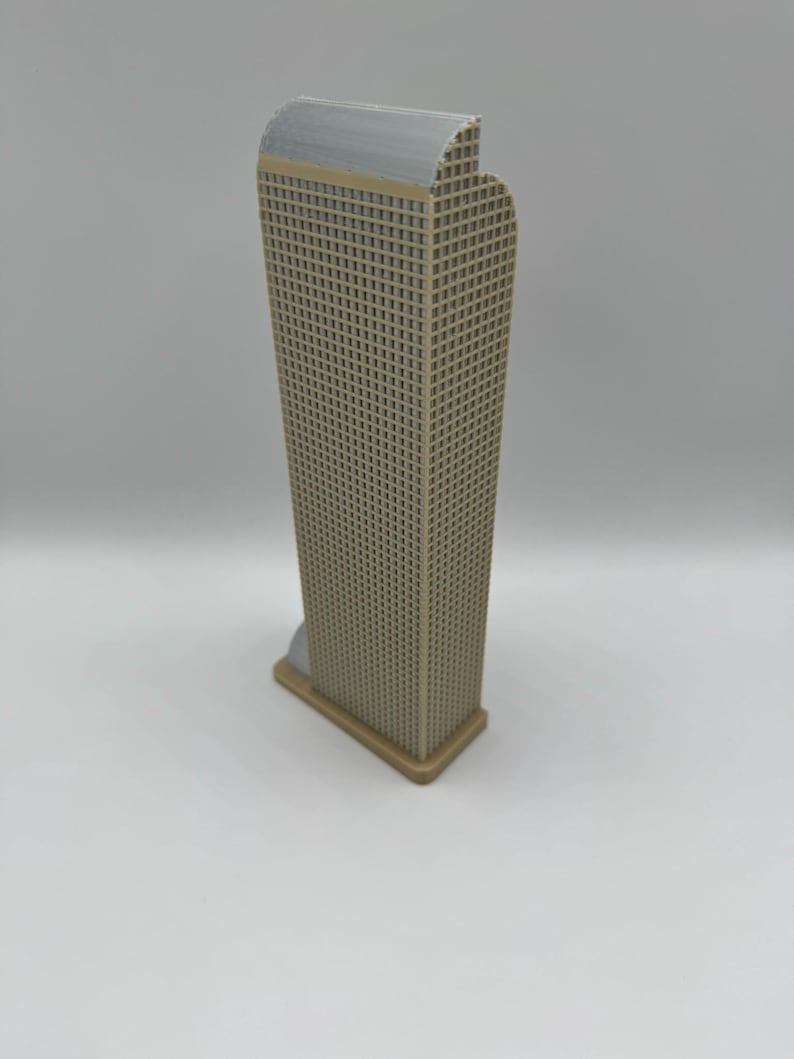 May include: A beige and gray miniature skyscraper model. The building has a rectangular shape with a grid pattern representing windows. The top of the building is gray, and the base is a lighter beige color. The model is standing on a white surface.