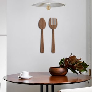 May include: A small, mid-century modern dining set in a minimalist kitchen.  The set includes a round wooden table with a dark brown top and black metal legs, and two light beige upholstered chairs with dark brown wooden legs.  On the wall above the table, there's a pair of wooden wall decor pieces shaped like a spoon and fork. A white pendant light hangs above the table.