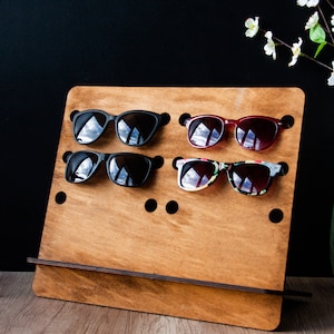 Puede incluir: Un expositor de madera con seis ranuras para mostrar gafas de sol. El expositor está hecho de madera marrón claro y tiene una base marrón oscuro. Hay tres pares de gafas de sol en el expositor, incluyendo gafas de sol negras, rojas y con estampado floral.