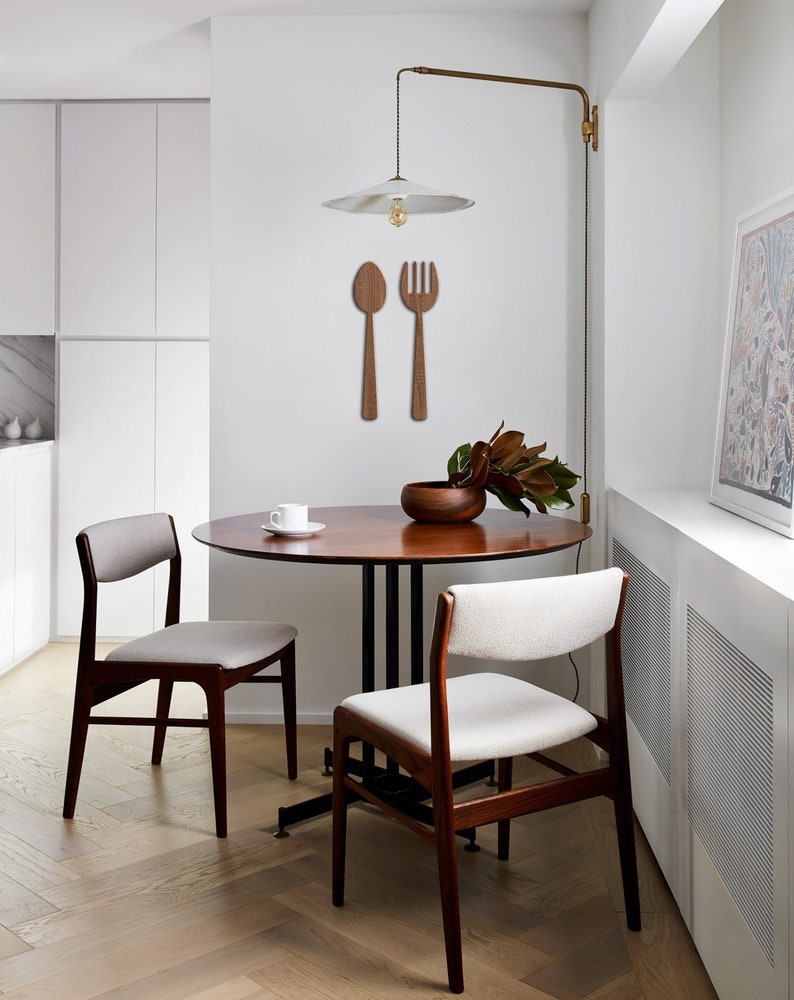 May include: A small, mid-century modern dining set in a minimalist kitchen.  The set includes a round wooden table with a dark brown top and black metal legs, and two light beige upholstered chairs with dark brown wooden legs.  On the wall above the table, there's a pair of wooden wall decor pieces shaped like a spoon and fork. A white pendant light hangs above the table.