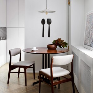 May include: A small, round wooden dining table with two mid-century modern style chairs. The chairs have light beige upholstery. A simple pendant light with a white shade hangs above the table.  Black wooden wall decor of a spoon and fork is mounted on the wall behind the table. A small bowl of greenery sits on the table.
