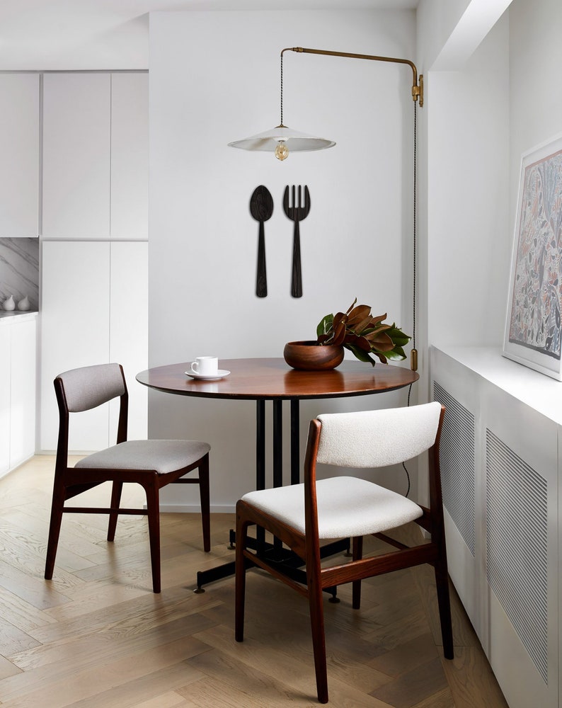 May include: A small, round wooden dining table with two mid-century modern style chairs. The chairs have light beige upholstery. A simple pendant light with a white shade hangs above the table.  Black wooden wall decor of a spoon and fork is mounted on the wall behind the table. A small bowl of greenery sits on the table.