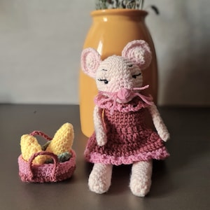 Può includere: Un topo rosa all'uncinetto che indossa un vestito bordeaux e tiene un cestino di banane gialle all'uncinetto. Il topo è seduto su una superficie grigia davanti a un vaso giallo con fiori secchi.