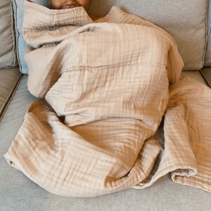 May include: A soft, light brown muslin blanket is folded and draped over a couch. The blanket is made of a lightweight, breathable fabric that is perfect for keeping warm on a cool day.