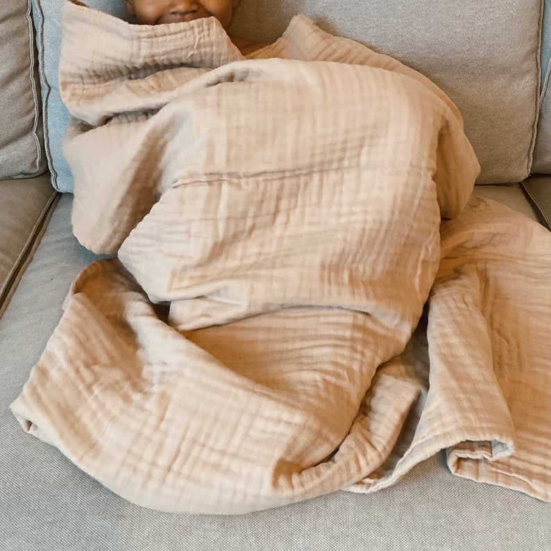 May include: A soft, light brown muslin blanket is folded and draped over a couch. The blanket is made of a lightweight, breathable fabric that is perfect for keeping warm on a cool day.