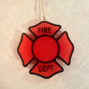 May include: A red Maltese cross-shaped ornament with a black border and black text reading FIRE DEPT. The ornament features a central circular section and four flared arms, suspended by a thin piece of twine attached to the top edge.