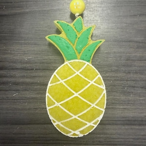 Op de afbeelding: Een hangend ornament in de vorm van een ananas met een geel getextureerd lichaam, een wit ruitpatroon en groene bladeren aan de bovenkant. Een enkele gele kraal is bevestigd aan het witte koord boven de ananas, ontworpen om op te hangen.
