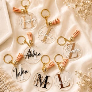 May include: Clear acrylic keyrings with gold-coloured hardware and peach-coloured tassels. Each keyring features a name and initial in various colours. The keyrings are personalised and suitable for gifts.
