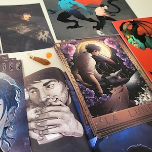 May include: A collection of art prints featuring illustrations of characters and creatures. The prints are on a white surface and include a comic book titled "Semi-Sacred".