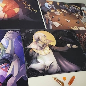 May include: Four different art prints featuring anime-style characters. The first print shows two characters in a romantic embrace. The second print shows a character with a white background and a yellow halo. The third print shows a character with a dark background and a red halo. The fourth print shows a character with a dark background and a red halo. The prints are all on a white surface.