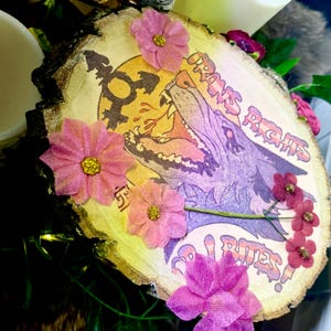 May include: A wooden round sign with a purple and pink wolf illustration and the text "Trans Rights Or I Bites!"  The sign is decorated with pink flowers.