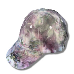 Op de afbeelding: Een tie-dye baseballpet met een groene, paarse en witte kleurstelling. De pet heeft een gebogen klep en witte stiksels.