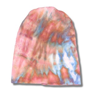 Op de afbeelding: Een tie-dye beanie in roze, blauwe en oranje tinten. De hoed heeft een zacht, getextureerd uiterlijk en een klassieke, afgeronde vorm. De kleuren vloeien samen in een wervelend patroon.