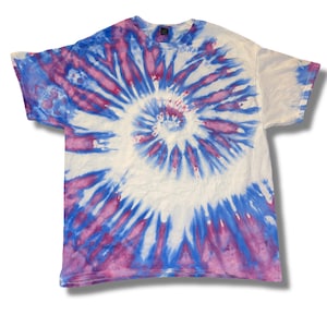 Op de afbeelding: Een wit t-shirt met een spiraalvormig tie-dye patroon in blauwe en paarse tinten. Het ontwerp straalt uit het midden, wat een opvallend effect creëert. Het shirt heeft korte mouwen en een ronde hals.