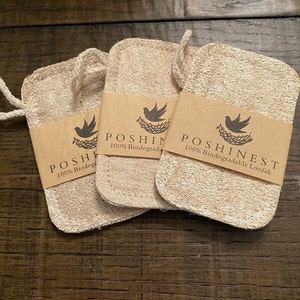 Puede incluir: Tres esponjas de baño de lufa beige con un lazo para colgar. Cada esponja tiene una banda de papel marrón con el texto "POSHINEST 100% Biodegradable Loofah" y un logotipo de un pájaro en un nido. Las esponjas están sobre una superficie de madera oscura.