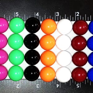 May include: A row of 10 colorful round beads, each bead is a different color: purple, pink, green, black, orange, white, maroon, blue, and green. The beads are arranged in a line on a white surface with a ruler behind them.