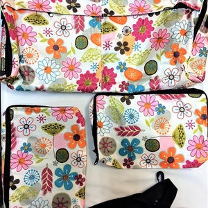 Op de afbeelding: Een set bloemenpatroontassen in verschillende maten. De tassen hebben een witte achtergrond met een kleurrijke print van bloemen in roze, oranje, blauw en groen. De set bevat een grote draagtas, een middelgrote tas en een kleinere pouch, allemaal met zwarte rand.