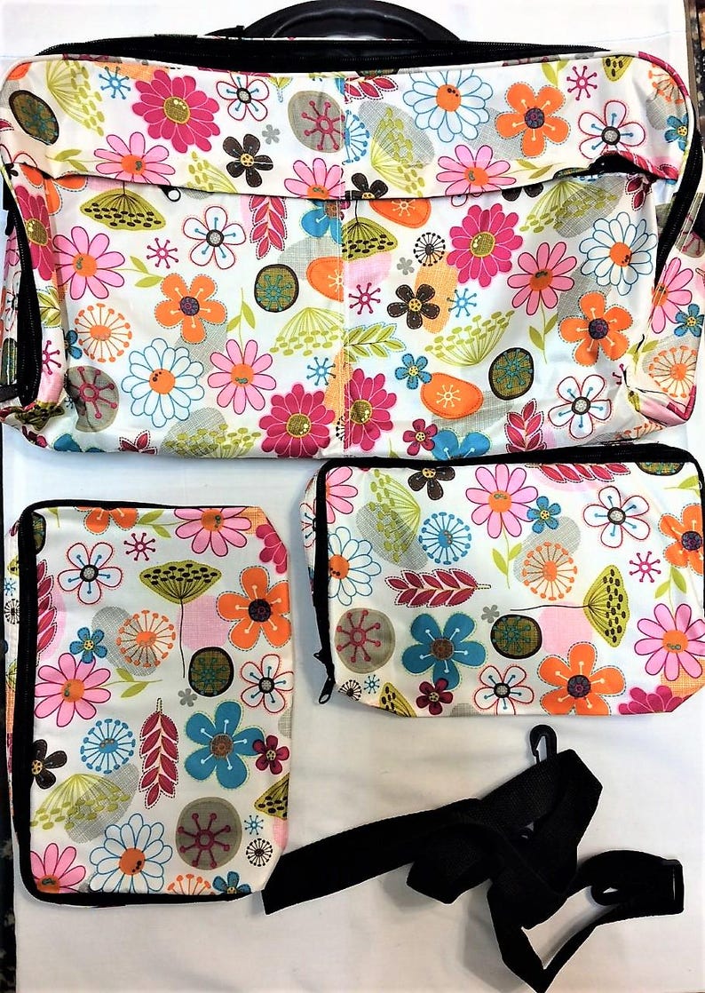 Op de afbeelding: Een set bloemenpatroontassen in verschillende maten. De tassen hebben een witte achtergrond met een kleurrijke print van bloemen in roze, oranje, blauw en groen. De set bevat een grote draagtas, een middelgrote tas en een kleinere pouch, allemaal met zwarte rand.