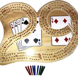 May include: A wooden cribbage board with a curved design, featuring a playing card display and scoring holes. The board includes a "START" and "FINISH" area, with a set of colored pegs for tracking scores. Cards show the 5 of hearts, spades, and diamonds.