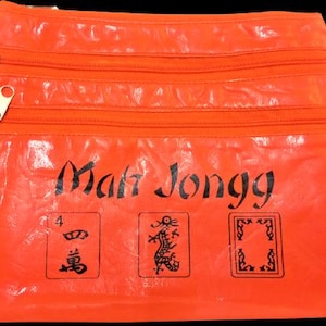 May include: Bright orange Mah Jongg pouch with three zippered compartments. The pouch features the text "Mah Jongg" in black, along with three illustrated tiles. Gold-colored zippers and pulls complete the design.