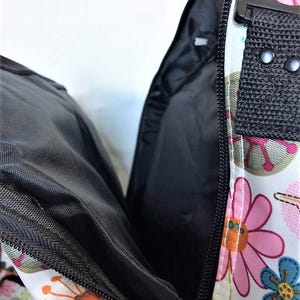 Op de afbeelding: Close-up van een rugzak met bloemenpatroon, met het open interieur. De tas heeft een witte basis met roze, oranje en blauwe bloempatronen. De binnenkant is zwart, met een zwarte rits en handvat. De tas is gedeeltelijk open.