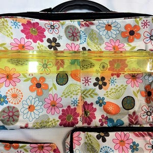 Op de afbeelding: Een totebag met bloemenpatroon met een zwart handvat en rits. De tas heeft een witte achtergrond met een kleurrijk scala aan bloemen in roze, oranje, blauw en bruin. Een doorzichtige, gele liniaal ligt over de bovenkant van de tas.