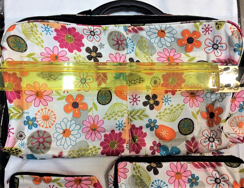 Op de afbeelding: Een totebag met bloemenpatroon met een zwart handvat en rits. De tas heeft een witte achtergrond met een kleurrijk scala aan bloemen in roze, oranje, blauw en bruin. Een doorzichtige, gele liniaal ligt over de bovenkant van de tas.