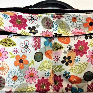 Op de afbeelding: Een tas met bloemenpatroon, zwarte handgreep en ritsen. Het ontwerp heeft een witte achtergrond versierd met roze, oranje, blauwe en bruine bloemen. De tas is ontworpen voor het dragen van spullen.