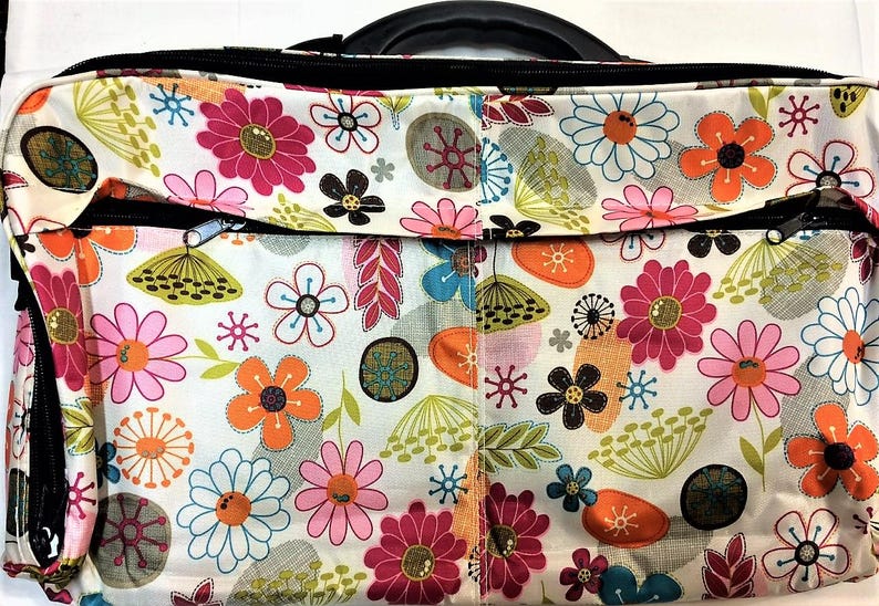 Op de afbeelding: Een tas met bloemenpatroon, zwarte handgreep en ritsen. Het ontwerp heeft een witte achtergrond versierd met roze, oranje, blauwe en bruine bloemen. De tas is ontworpen voor het dragen van spullen.