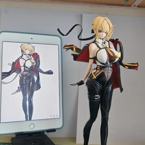 Peut inclure: Figurine détaillée d'un personnage d'anime féminin aux cheveux blonds, vêtue d'une tenue noire et blanche avec une cape rouge. La figurine tient un petit couteau. Une tablette affiche une image similaire.