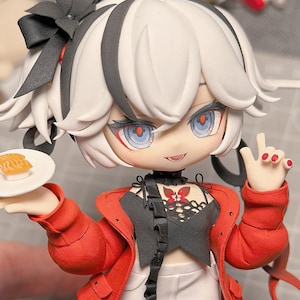 May include: A detailed figurine of a character with white hair, black accents, and blue eyes. The character wears a red jacket, black top, and white shorts, holding a plate with food. The figurine has a bow in its hair and is making a peace sign.