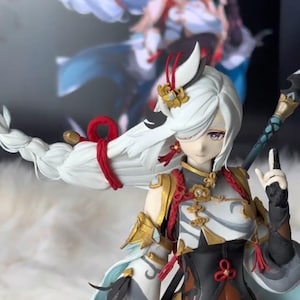 May include: A detailed figurine of a character with long white hair, adorned with red accents and gold detailing. The figure wears a black and white outfit with intricate patterns. A blurred image of a similar character is in the background.