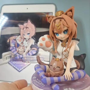 May include: A detailed figurine of a cat-eared anime character with blue eyes, wearing a white and brown outfit. The figure is holding a paw-shaped object and is seated on a purple base with a cat. A smaller version of the figure is visible in the background.
