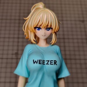 Może przedstawiać: Kolekcjonerska figurka w stylu anime przedstawiająca postać z blond włosami związanymi w kucyk i jasnoniebieskimi oczami. Figurka ma na sobie jasnoniebieski t-shirt z napisem WEEZER wydrukowanym czarnymi wielkimi literami na klatce piersiowej.