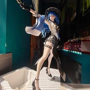 Może przedstawiać: Stojąca figurka w stylu anime przedstawiająca postać z długimi niebieskimi włosami, w kapeluszu z szerokim rondem, biało-czarnym stroju z asymetryczną spódnicą i czarnych butach na wysokim obcasie. Postać trzyma w górze mały pistolet.
