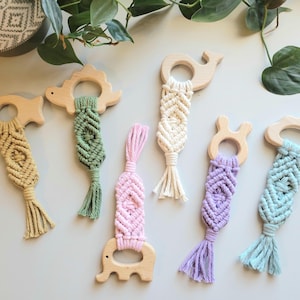 May include: Six wooden teethers with macrame tassels in various colors, including green, pink, purple, and blue. The teethers are shaped like a dinosaur, whale, elephant, bunny, and cloud.