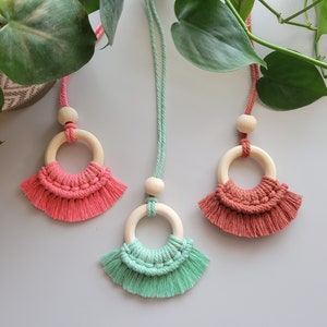 May include: Three macrame necklaces with wooden rings and tassels. The necklaces are in shades of pink, green, and brown.