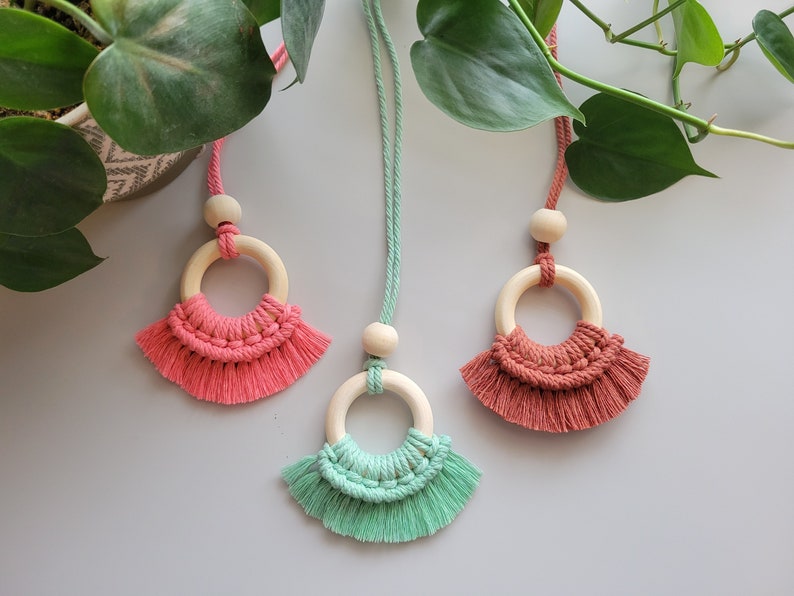 May include: Three macrame necklaces with wooden rings and tassels. The necklaces are in shades of pink, green, and brown.