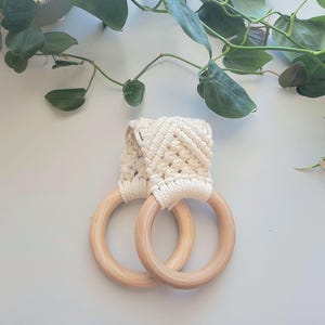 May include: A pair of wooden towel rings with a cream-colored macrame holder. The rings are made of light-colored wood and are connected by the woven macrame. Green leafy vines are in the background.