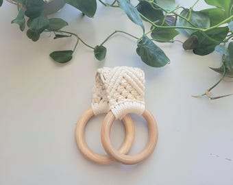 Puede incluir: Un par de aros de toalla de madera con un soporte de macramé color crema. Los aros son de madera de color claro y están conectados por el macramé tejido. En el fondo hay vides verdes y frondosas.