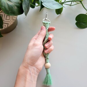 May include: A light green macrame keychain with a wooden bead and a tassel. The keychain is held in a hand.