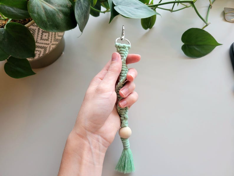 May include: A light green macrame keychain with a wooden bead and a tassel. The keychain is held in a hand.