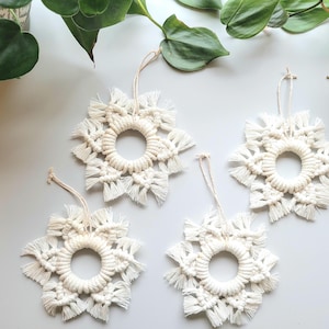 May include: Four off-white macrame snowflake ornaments with hanging loops. Each ornament has a circular center and radiating fringe, resembling a snowflake. The ornaments are displayed on a light gray surface with green foliage.