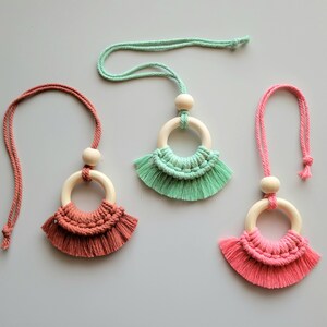 May include: Three macrame necklaces with wooden rings and tassels. The necklaces are in shades of pink, green, and coral.