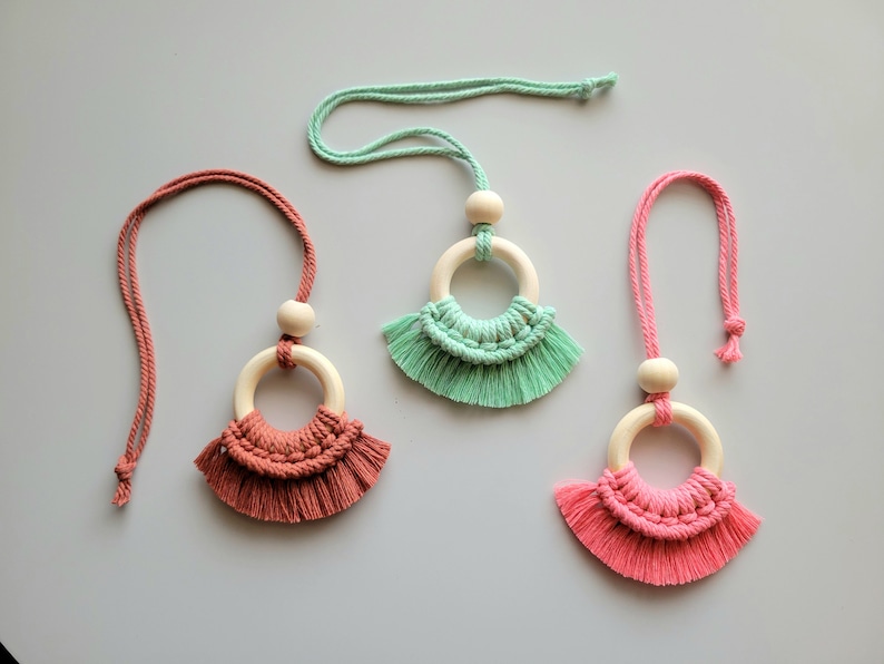 May include: Three macrame necklaces with wooden rings and tassels. The necklaces are in shades of pink, green, and coral.