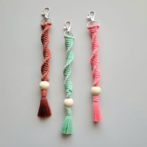 May include: Three handmade keychains with a wooden bead and tassel. The keychains are made with a twisted cord in pink, green, and brown.