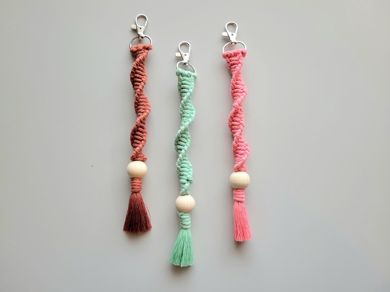 May include: Three handmade keychains with a wooden bead and tassel. The keychains are made with a twisted cord in pink, green, and brown.