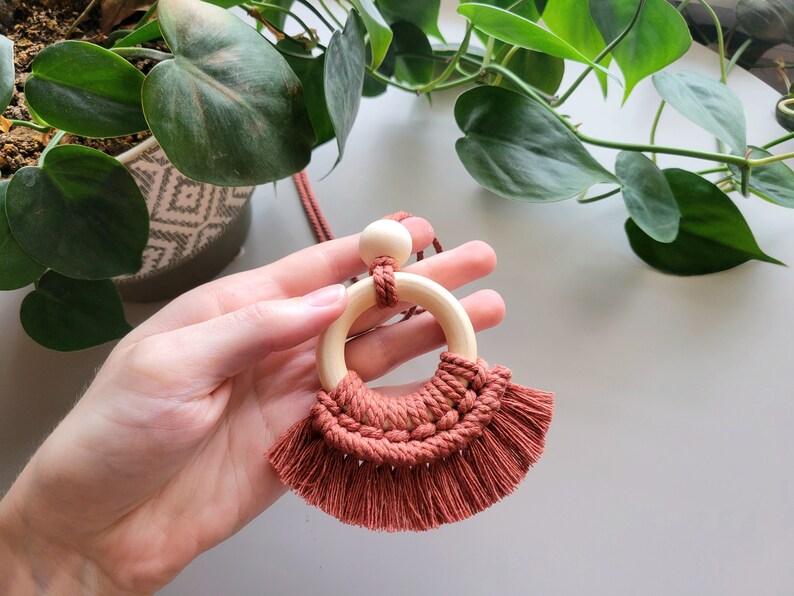 May include: A terracotta macrame necklace with a wooden ring and bead. The necklace has a fringe detail.