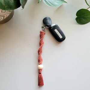 May include: A terracotta-coloured macrame keychain with a white bead and a tassel. The keychain is attached to a black car key.