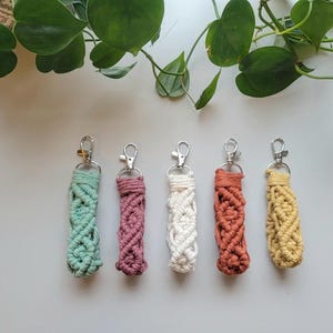 May include: Five colorful macrame keychains with silver key rings. The keychains are made with a variety of colors, including green, pink, white, orange, and yellow.